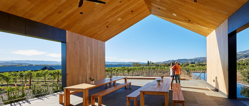 A couple standing under a wooden roof patio looking out into the vineyards and hills in the distance.