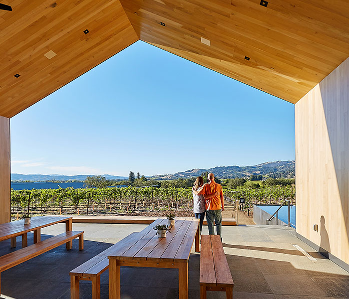 A couple standing under a wooden roof patio looking out into the vineyards and hills in the distance.