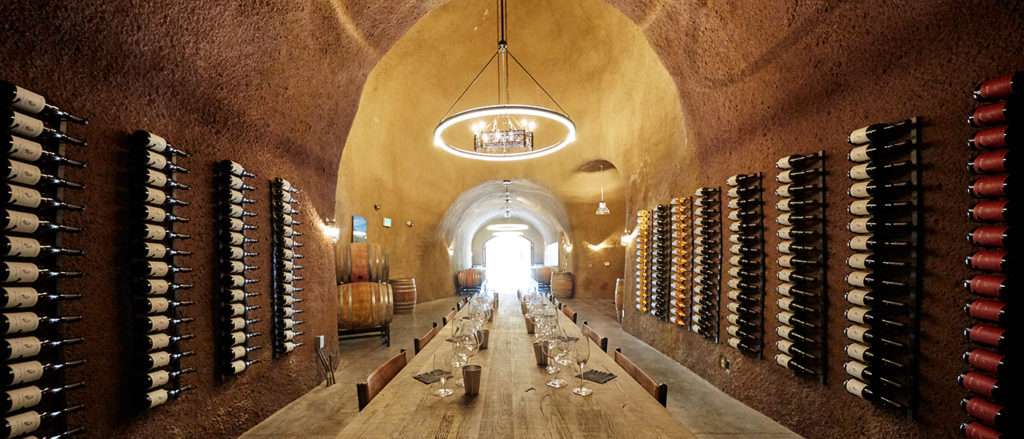 Long Wooden Table set for wine tasting surrounded by wine in rack displays on the wall of a wine cave.
