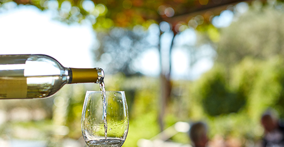 A bottle of white wine being poured into a wine glass, garden in the background.
