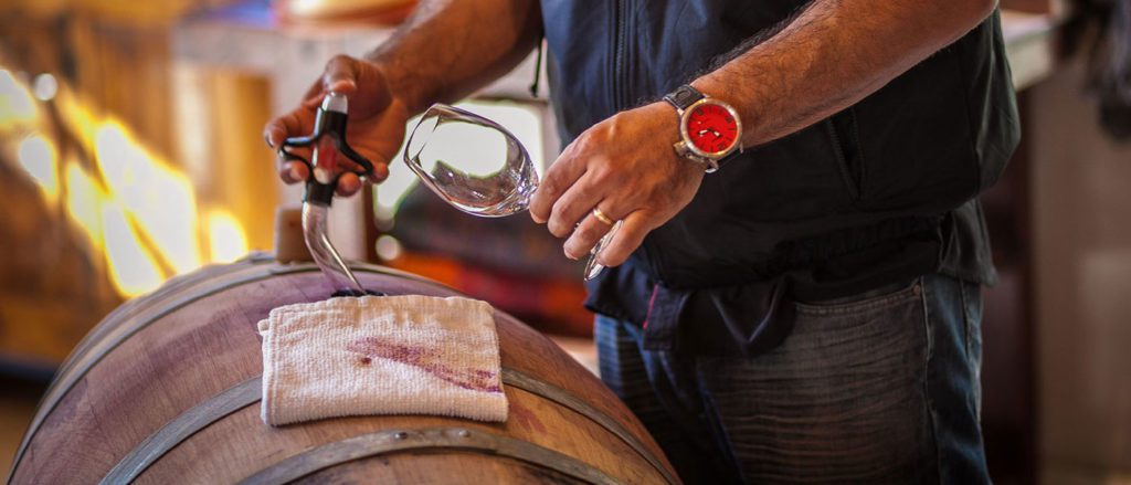 Winemaker pulling wine from a barrel with a wine thief while holding a wine glass.