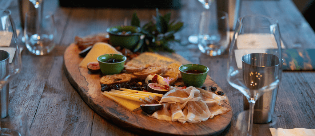 An oval wood board with crackers, various cheeses, jams, dried fruit and prosciutto on a wood table with wine glasses