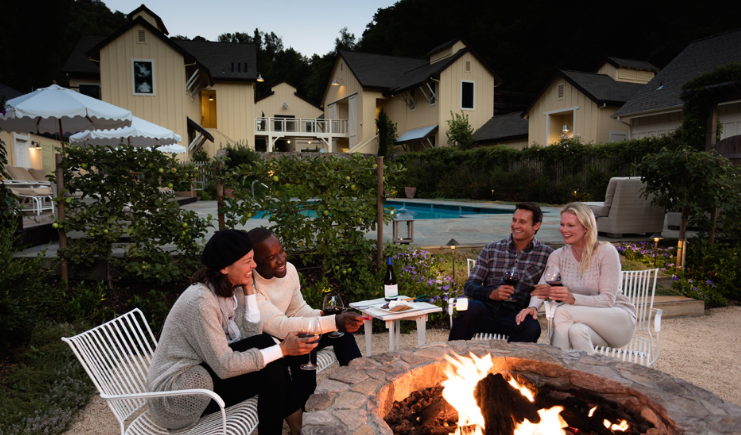 Two couples sitting around a fire place near a pool drinking wine and laughing