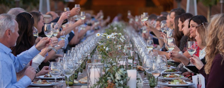 Long table set for an outdoor dinner with guests toasting with their wine glasses in the air.