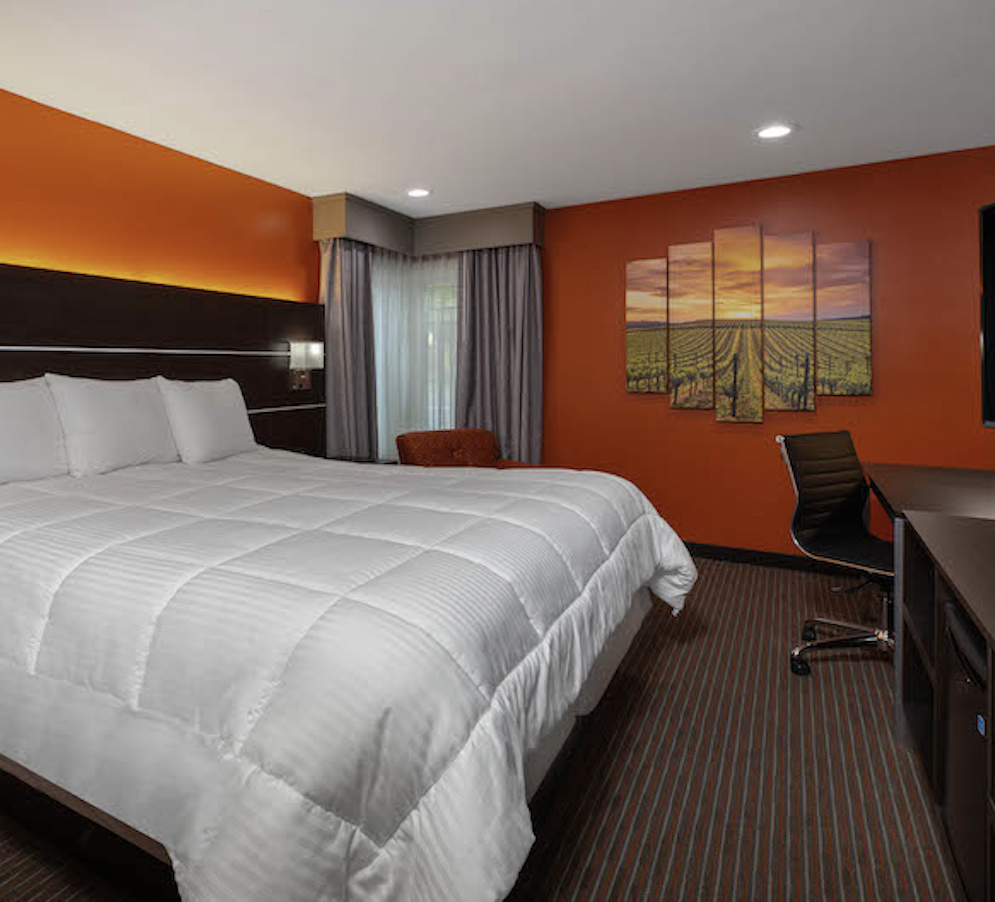 White duvet on a queen size bed, dark furniture, grey drapes and orange walls with picture of a vineyard scene.