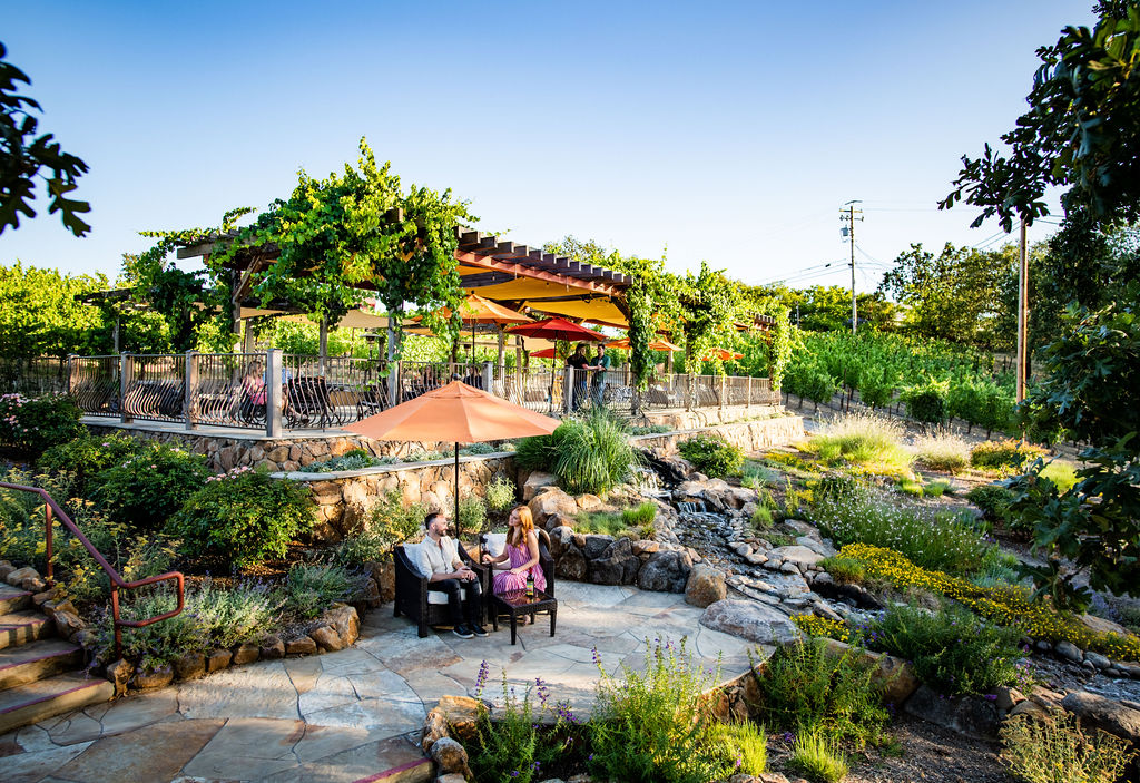 A couple shares wine under an orange umbrella in front of pergola covered outdoor landscaped patio.