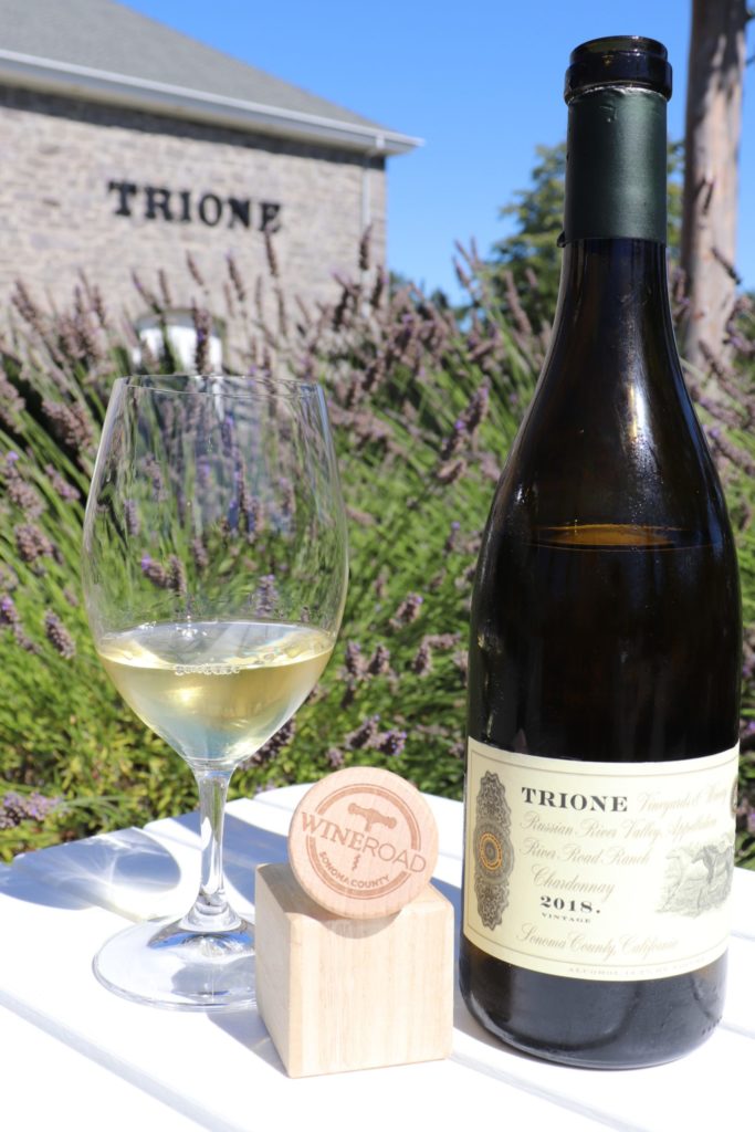 A glass and bottle of Trione chardonnay with a lavender bush and the Old Stone Building behind.