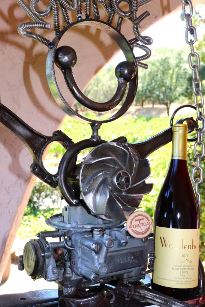 A bottle of Woodenhead Pinot Noir nestled in a hanging metal piece of art with the vineyard in the back.