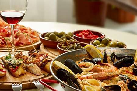 a partial glass of red wine among plates of appetizer foods and a paella pan filled with seafood.